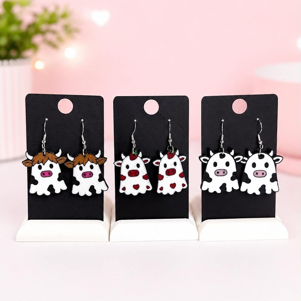 Three pairs of cow-shaped dangle earrings displayed on black card holders against a soft pink background with blurred lights and a plant.