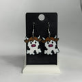 A pair of cow-shaped dangling earrings displayed on a black card stand against a light gray background. The earrings feature white bodies with black spots, brown hair, and pink snouts, suspended from silver hooks.