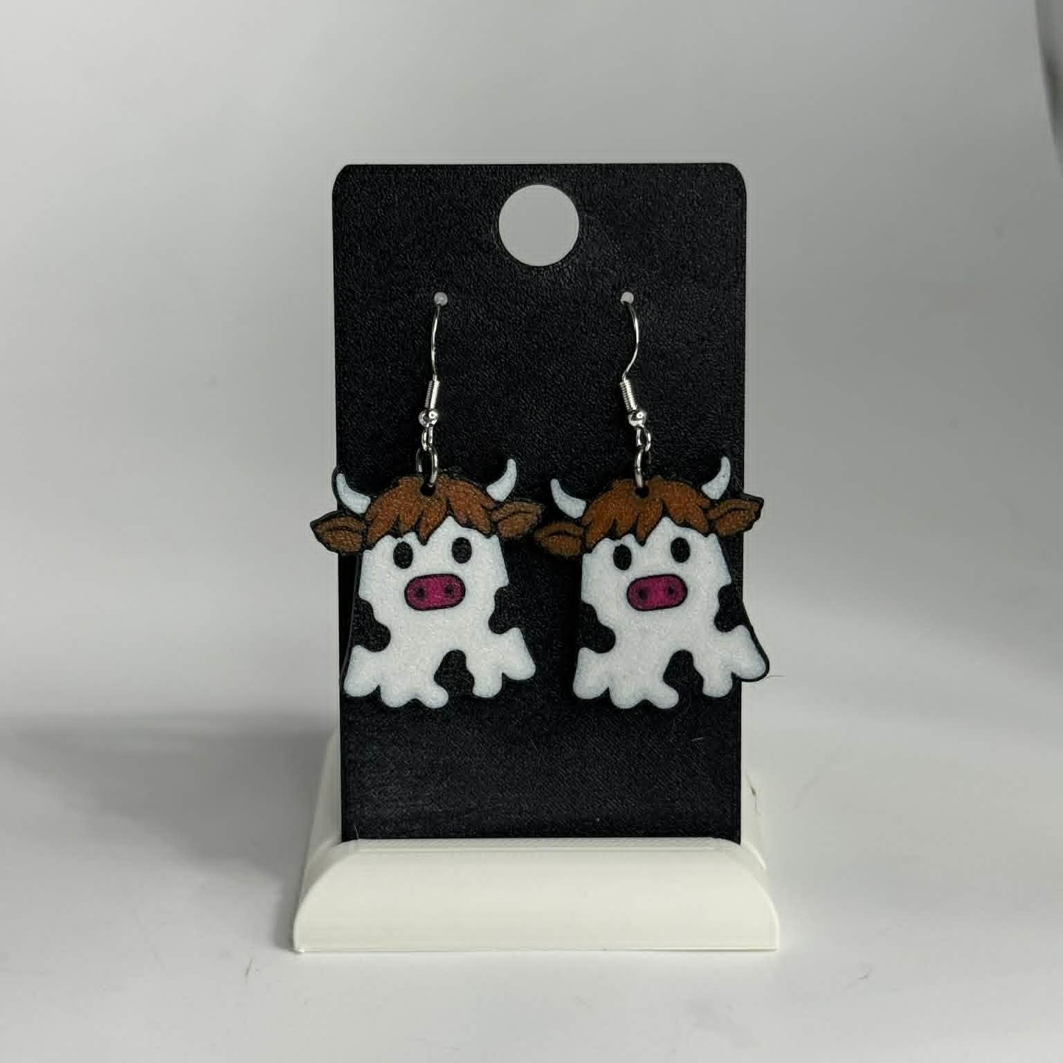 A pair of cow-shaped dangling earrings displayed on a black card stand against a light gray background. The earrings feature white bodies with black spots, brown hair, and pink snouts, suspended from silver hooks.