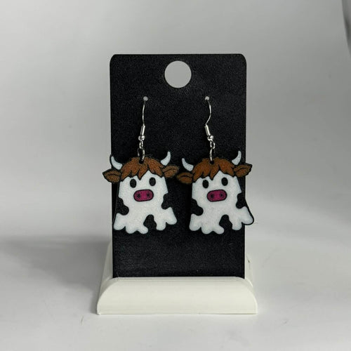 A pair of cow-shaped dangling earrings displayed on a black card stand against a light gray background. The earrings feature white bodies with black spots, brown hair, and pink snouts, suspended from silver hooks.
