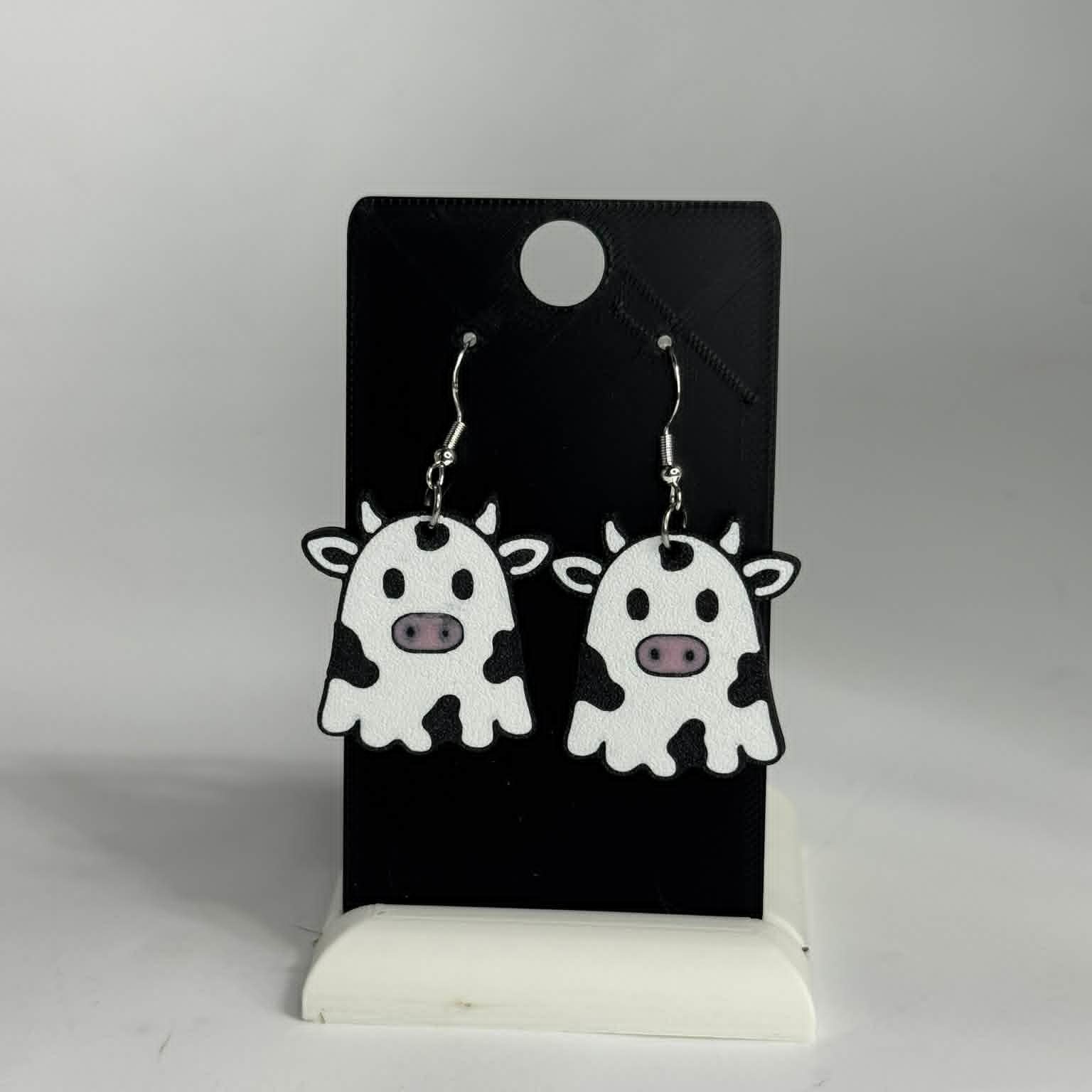 A pair of cow-shaped dangling earrings displayed on a black card stand against a light gray background. The earrings are white with black spots and small pink noses, hanging from silver hooks.