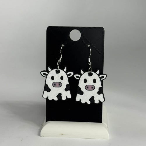 A pair of cow-shaped dangling earrings displayed on a black card stand against a light gray background. The earrings are white with black spots and small pink noses, hanging from silver hooks.
