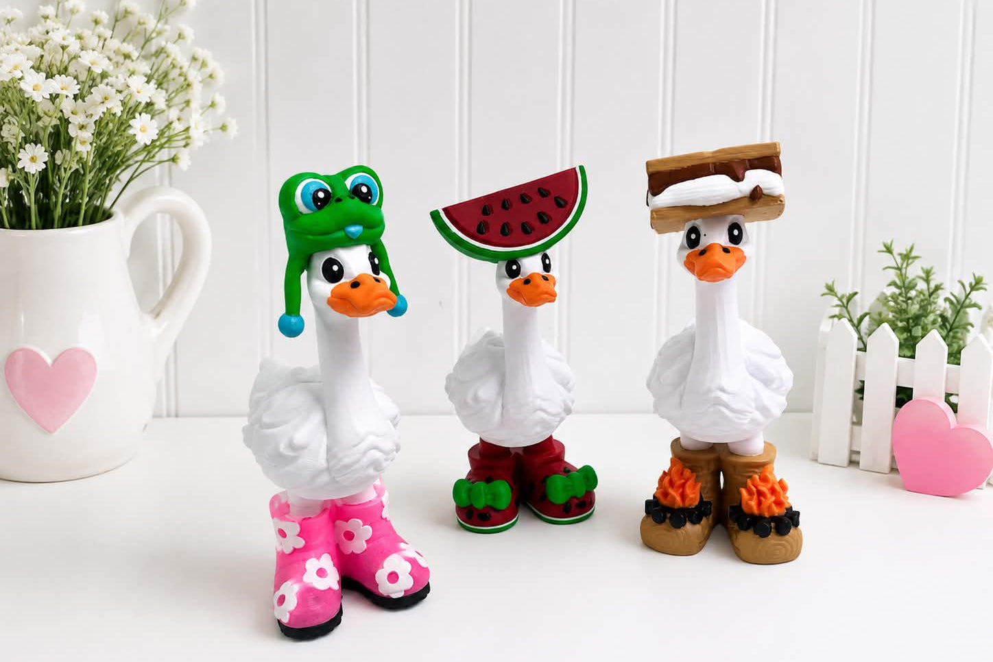 Three whimsical goose figurines styled in playful outfits: one in pink floral rain boots with a green frog hat, one in red and green watermelon boots, and one in brown and black boots with a sandwich hat. They are displayed on a white surface with decorative props including a white pitcher of flowers, a small plant, and pink heart accents.