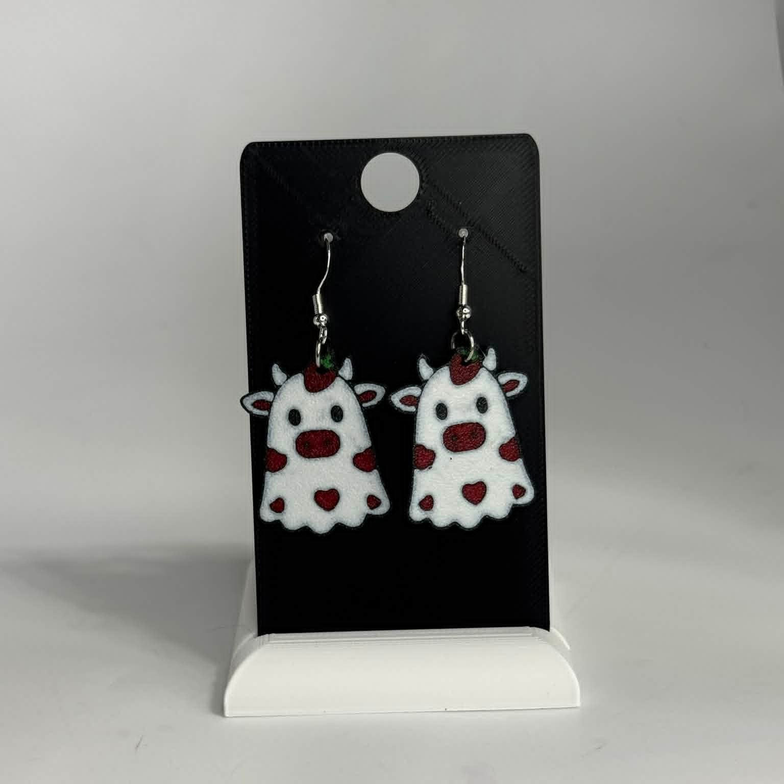 A pair of white cow-shaped dangle earrings with red hearts and spots, displayed on a black card stand against a light gray background.